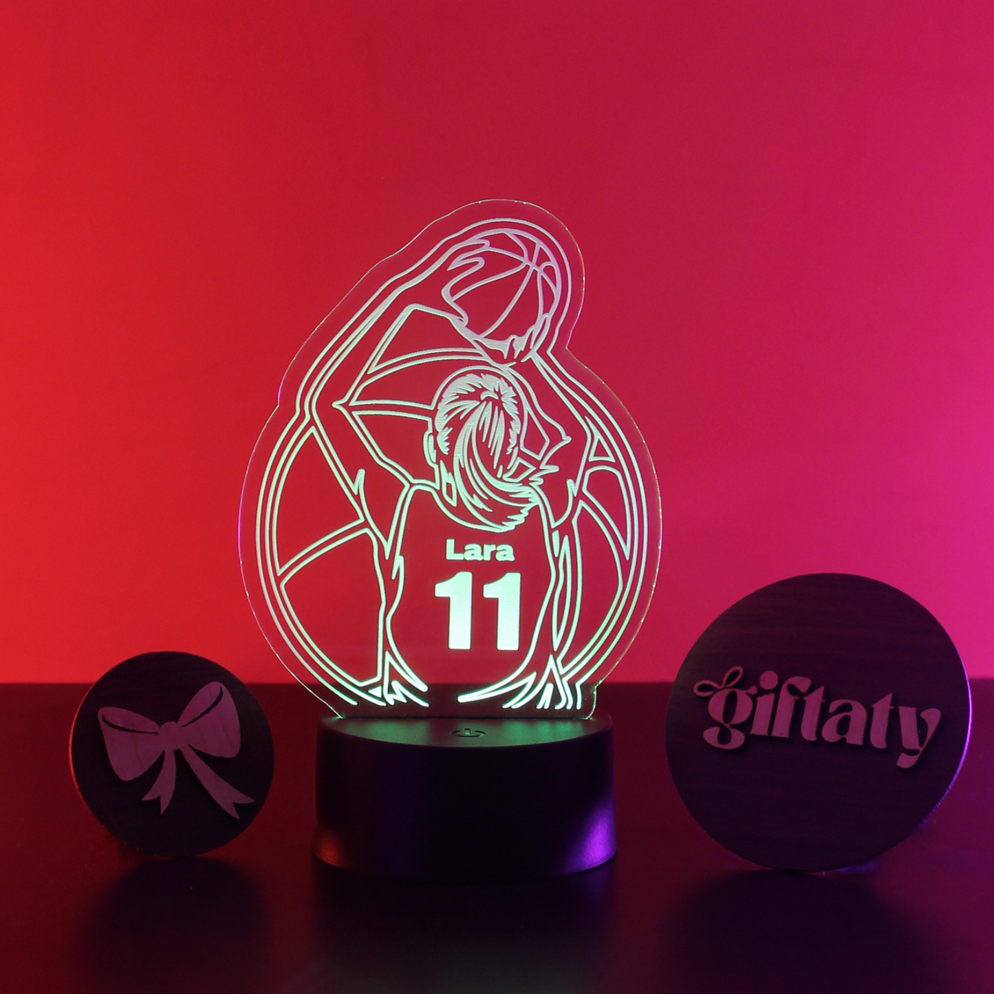 A personalized 3D optical illusion night light featuring a basketball player silhouette with the name 'Lara' and number '11' on the illuminated acrylic panel, set on a black base. The scene is flanked by two round black wooden tokens engraved with a bow and the name 'GiIaty', all set against a vibrant pink and red gradient background.