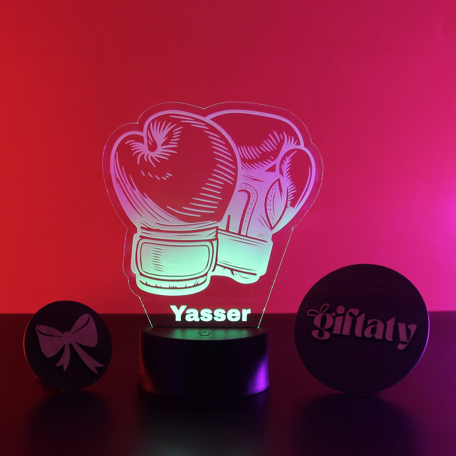 A glowing acrylic 3D lamp shaped like a pair of boxing gloves on a black cylindrical base, with the name 'Yasser' illuminated in the center. Two round black gift tags are placed beside it on a dark surface against a red-to-pink gradient background.