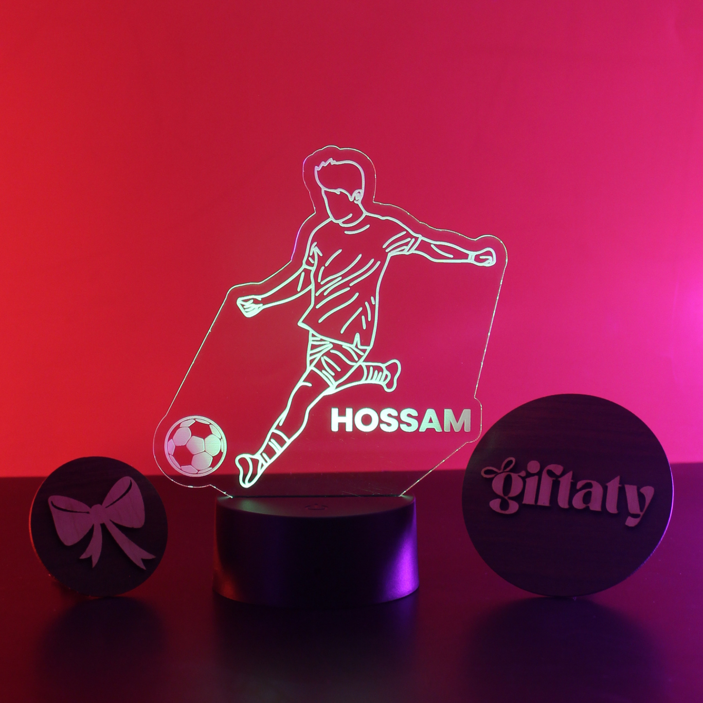 A personalized LED acrylic night light featuring a soccer player silhouette, a soccer ball, and the name 'HOSSAM' in the center, displayed on a black cylindrical base. Two round black gift boxes with pink laser-engraved designs are positioned on either side on a dark surface against a vibrant pink and red gradient background.