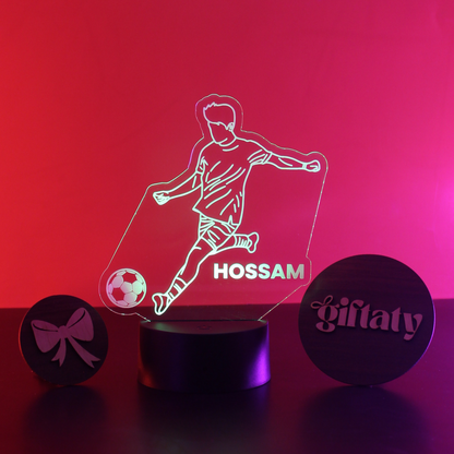 A personalized LED acrylic night light featuring a soccer player silhouette, a soccer ball, and the name 'HOSSAM' in the center, displayed on a black cylindrical base. Two round black gift boxes with pink laser-engraved designs are positioned on either side on a dark surface against a vibrant pink and red gradient background.