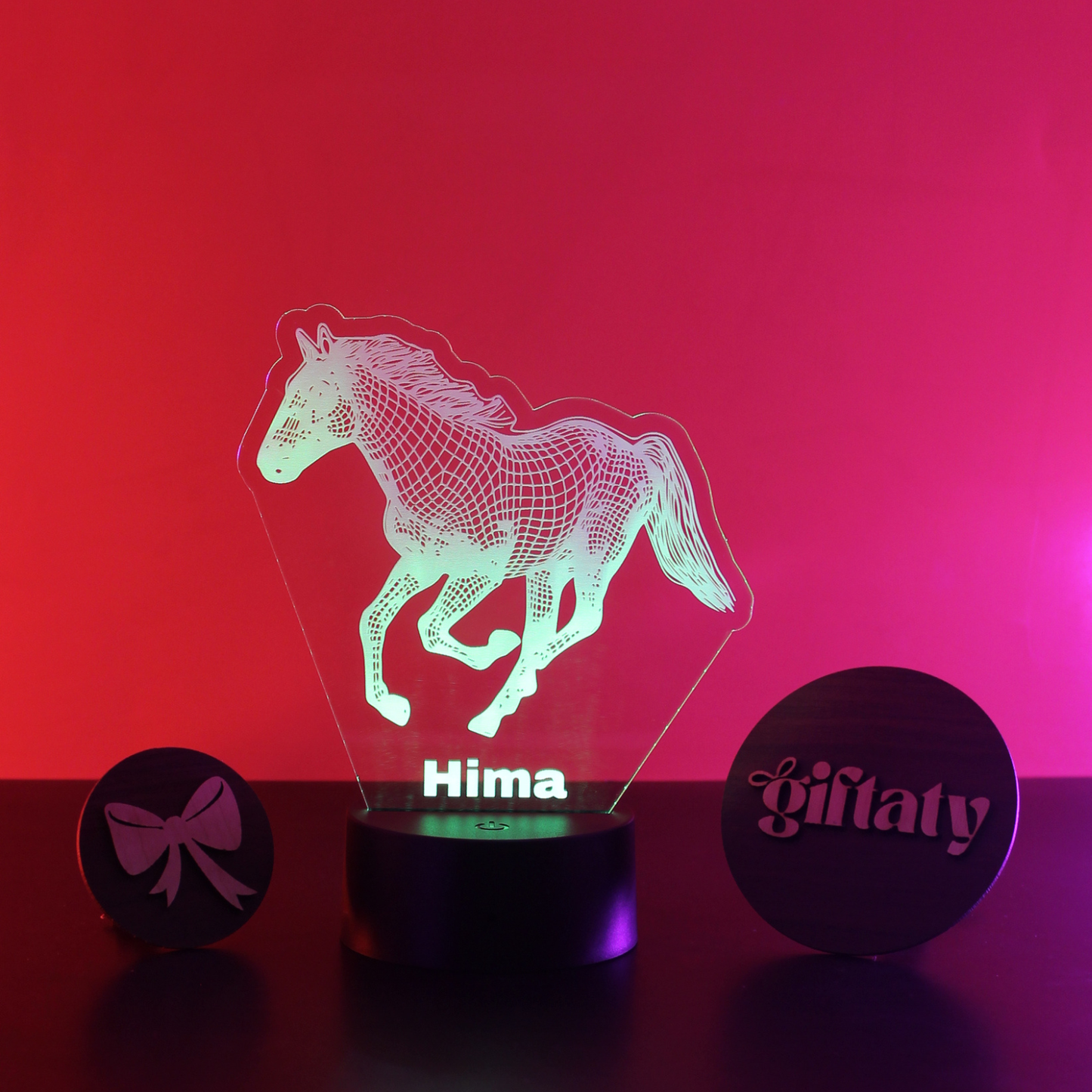 A 3D optical illusion lamp showing a running horse silhouette with the name 'Hima' on the front, displayed on a black base with two round black nameplates on either side against a red and pink gradient background.