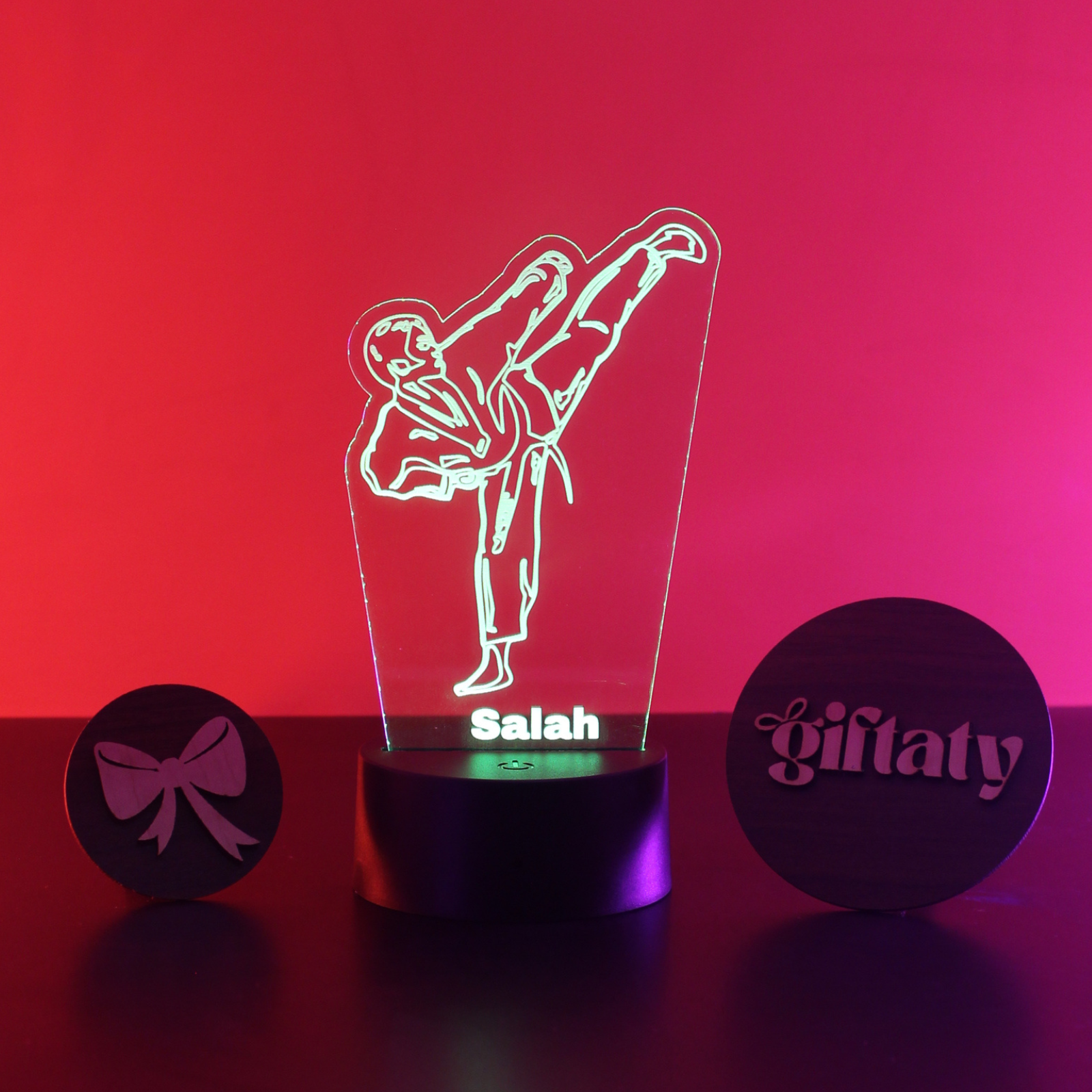 A personalized 3D acrylic LED night light featuring a martial artist silhouette in a high kick, with the name 'Salah' illuminated on the base. The light is displayed on a dark surface against a vibrant pink and red gradient background, flanked by two round black decorative discs engraved with 'giftaty' and a bow.