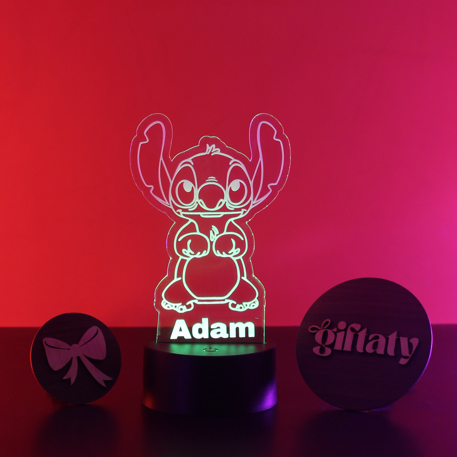 A personalized acrylic Disney Stitch night light on a black base, illuminated with a green outline and the name 'Adam' at the bottom. Two round black gift boxes are placed on either side on a red and pink gradient background.