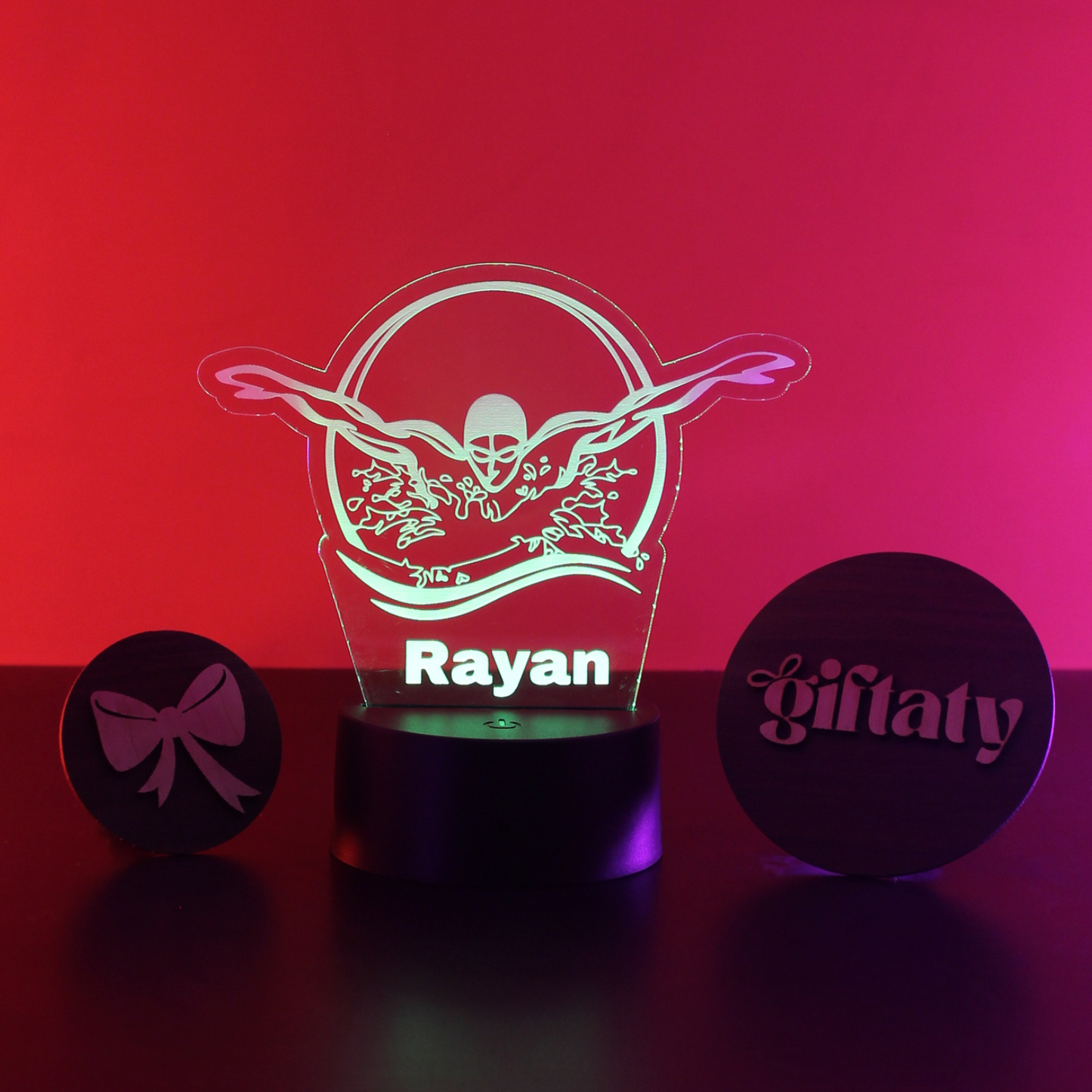 A personalized acrylic 3D optical illusion night light shaped like a winged skull with the name 'Rayan' illuminated in the center, displayed on a black base against a red-to-pink gradient background. Two round black gift tags are placed beside it, one with a bow icon and the other with the name 'giftaty'.