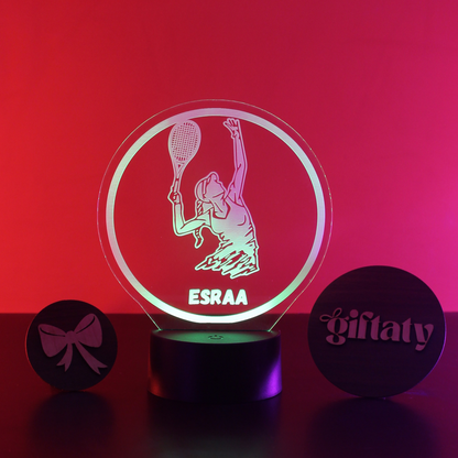 A personalized acrylic LED night light on a black base, featuring a tennis player silhouette and the name 'ESRAA' illuminated in green and pink. Two small round black gift tags are placed on either side on a red-lit background.