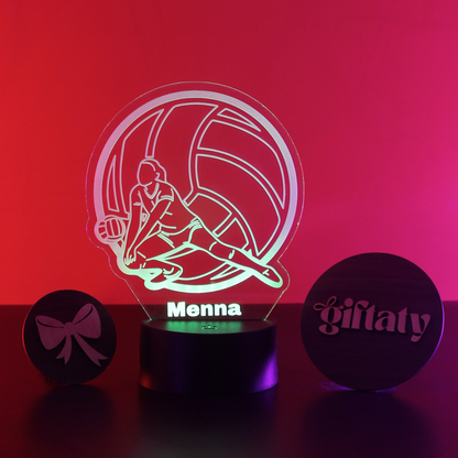 A personalized 3D acrylic LED night light showing a volleyball player silhouette with the name 'Menna' on the base, flanked by two engraved wooden keepsakes on a dark surface against a red-to-pink gradient background.